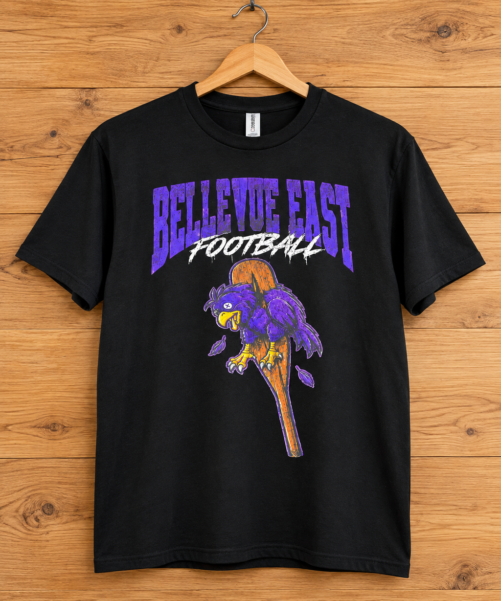 A black short-sleeve graphic t-shirt hanging on a wooden hanger against a wood plank wall. The shirt features a purple and white 'BELLEVOE EAST FOOTBALL' graphic with a purple and gold football mascot illustration.